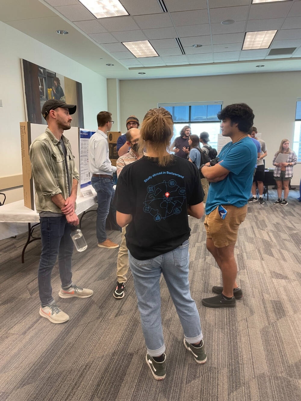 A group of people stand in a carpeted room in front of poster boards, chatting casually in what appears to be a university or conference poster session. One person wears a black T-shirt with “Study abroad in Switzerland” and a map graphic.